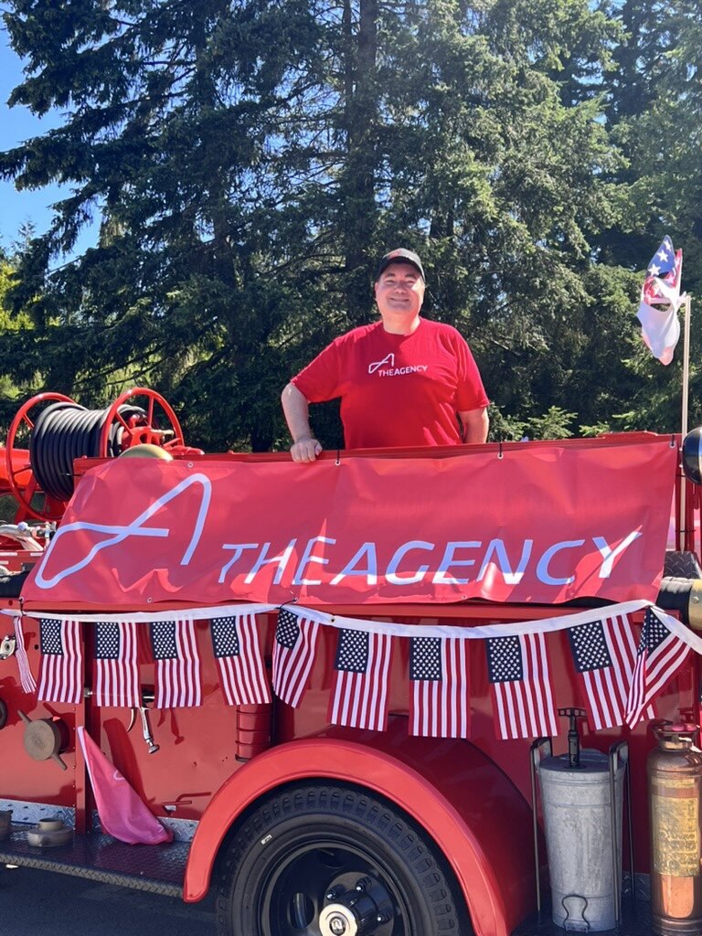Raymond in a red The Agency T-shirt aboard a vintage red fire truck draped with The Agency banner and American flag bunting, Bainbridge Island Grand Old Fourth parade.
