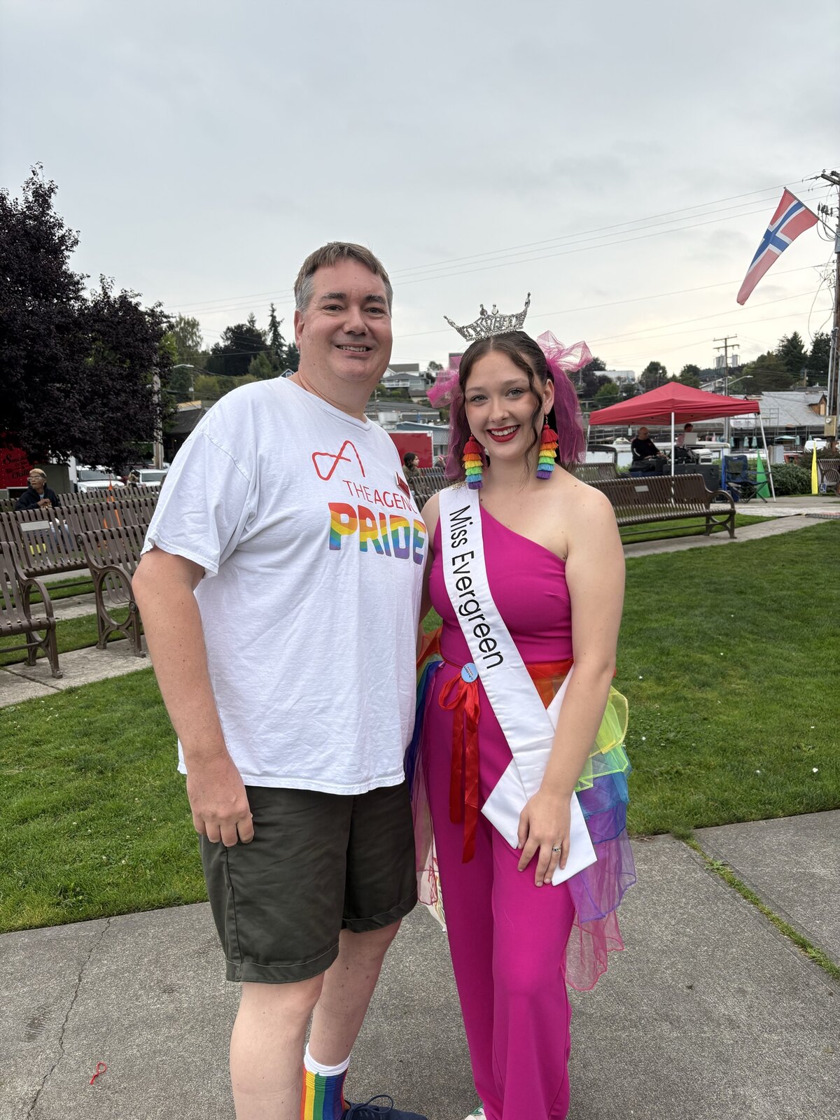 Raymond in a white The Agency Pride T-shirt standing next to Miss Evergreen wearing a rainbow tutu and crown at Poulsbo Pride.