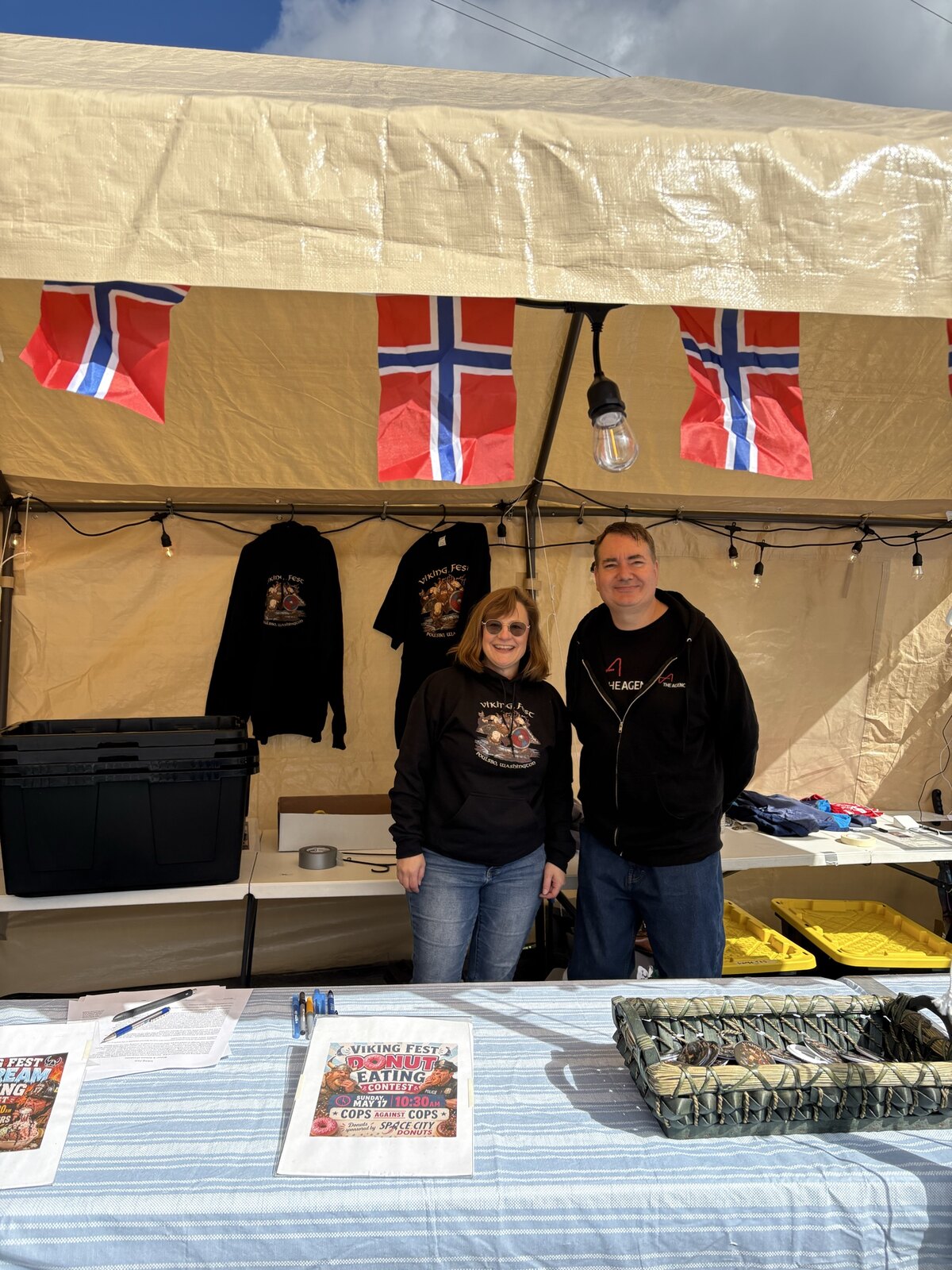 Raymond in a red The Agency hoodie next to Debbie Nazarino-Skolrud at the Viking Fest Donut Eating Contest booth in Poulsbo, Norwegian flags overhead.