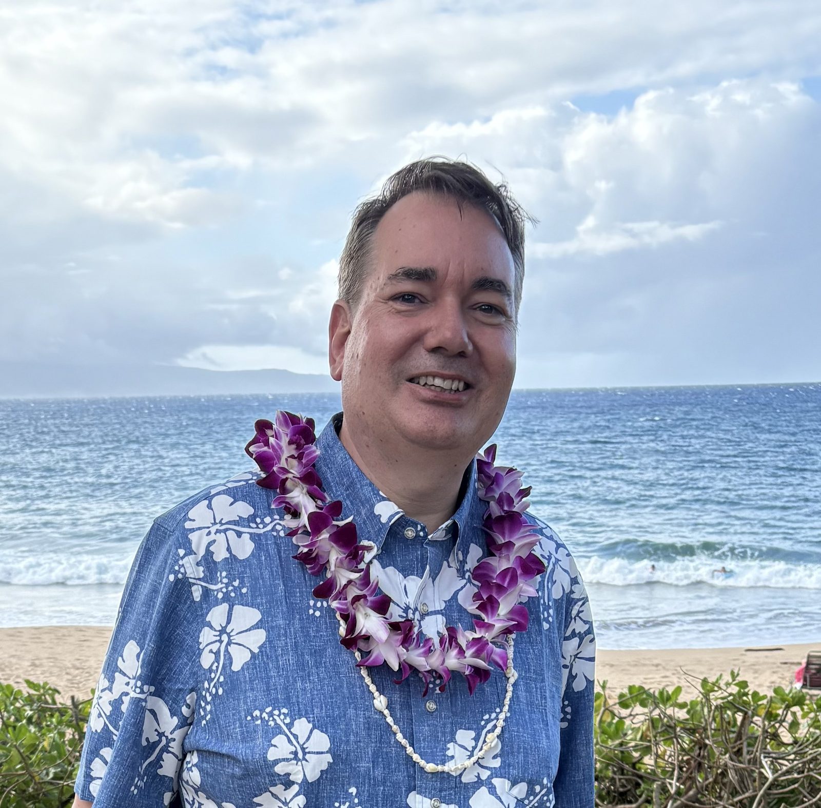 Raymond wearing a purple orchid lei on a Maui beach, ocean and clouds in the background.