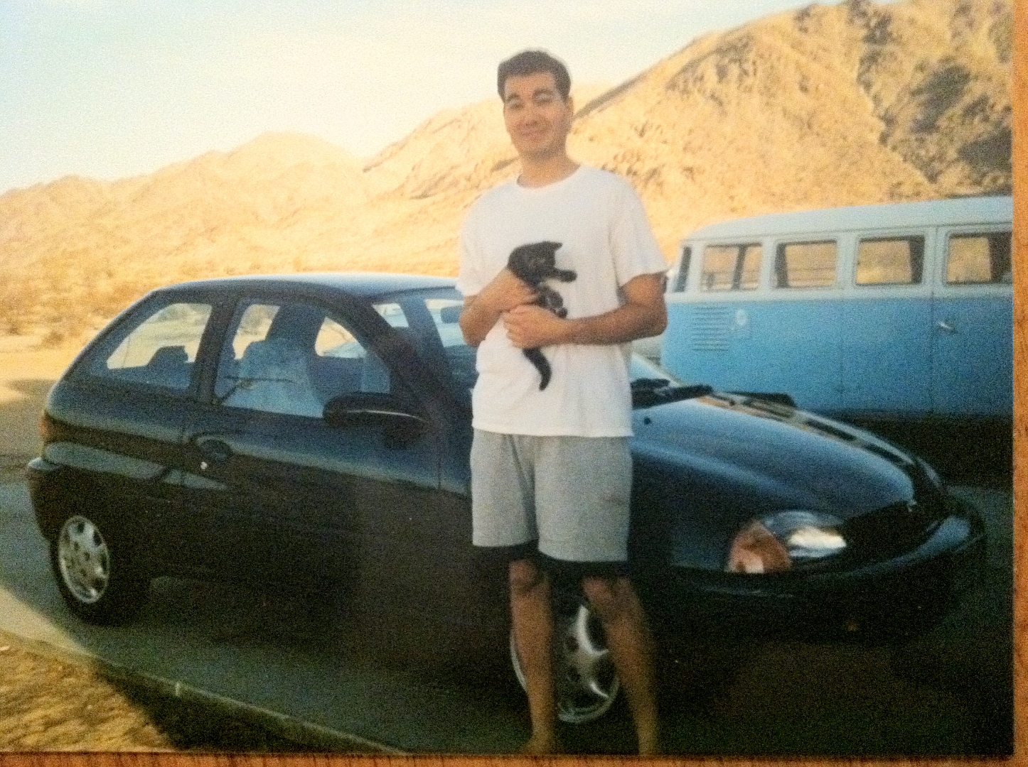 A younger Raymond in the desert beside a small black hatchback and a vintage blue Volkswagen bus, holding a tiny black kitten.
