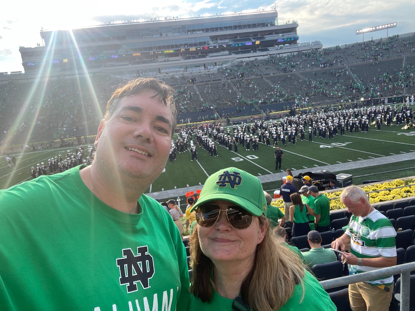 Raymond and his wife at a Notre Dame football game in Notre Dame Stadium, wearing green Fighting Irish gear with the marching band on the field.