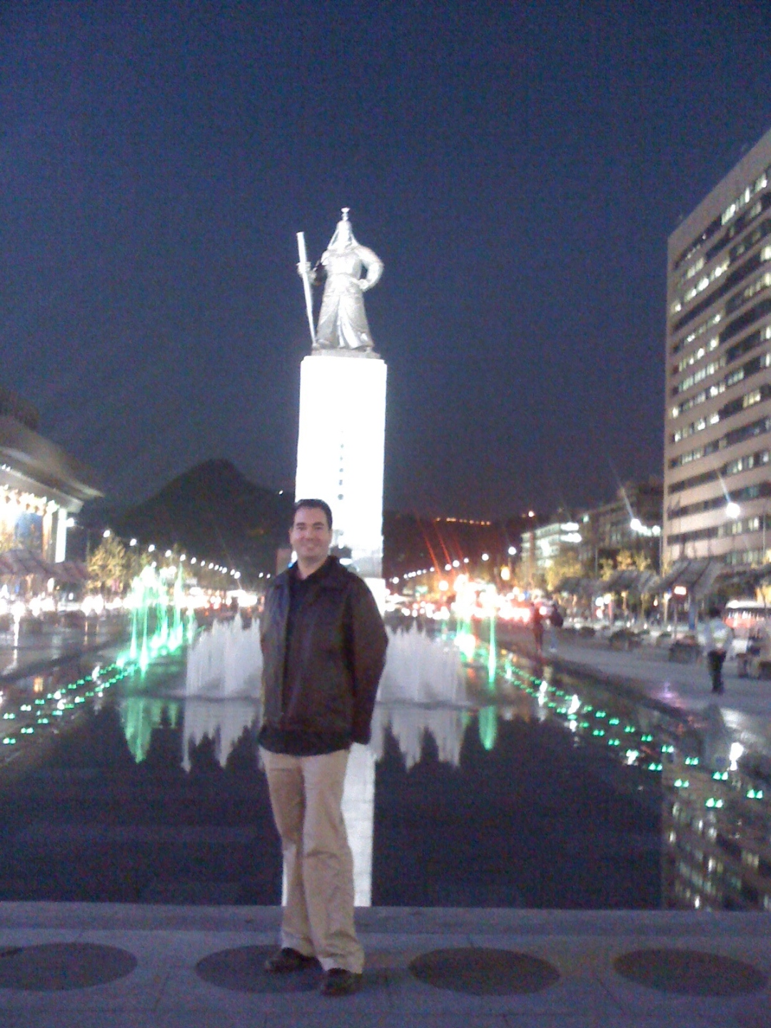 Raymond at night in Gwanghwamun Plaza in Seoul, lit fountains and the Admiral Yi Sun-sin statue behind him.