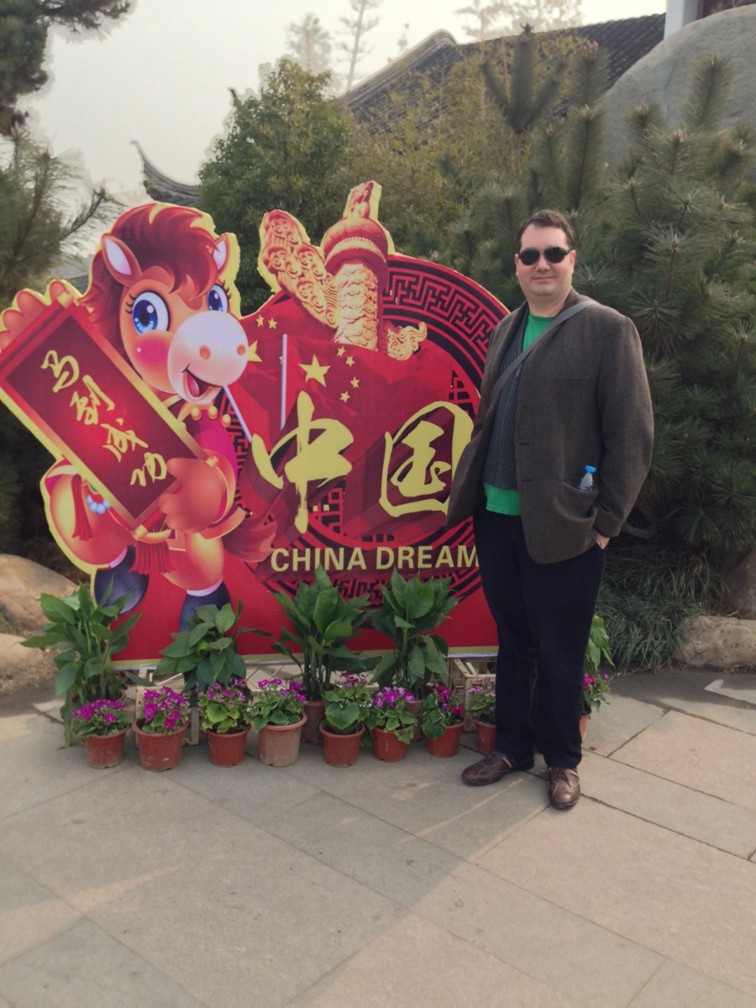 Raymond on a stone walkway in a Chinese garden next to a red China Dream Lunar New Year horse display, surrounded by potted flowers.