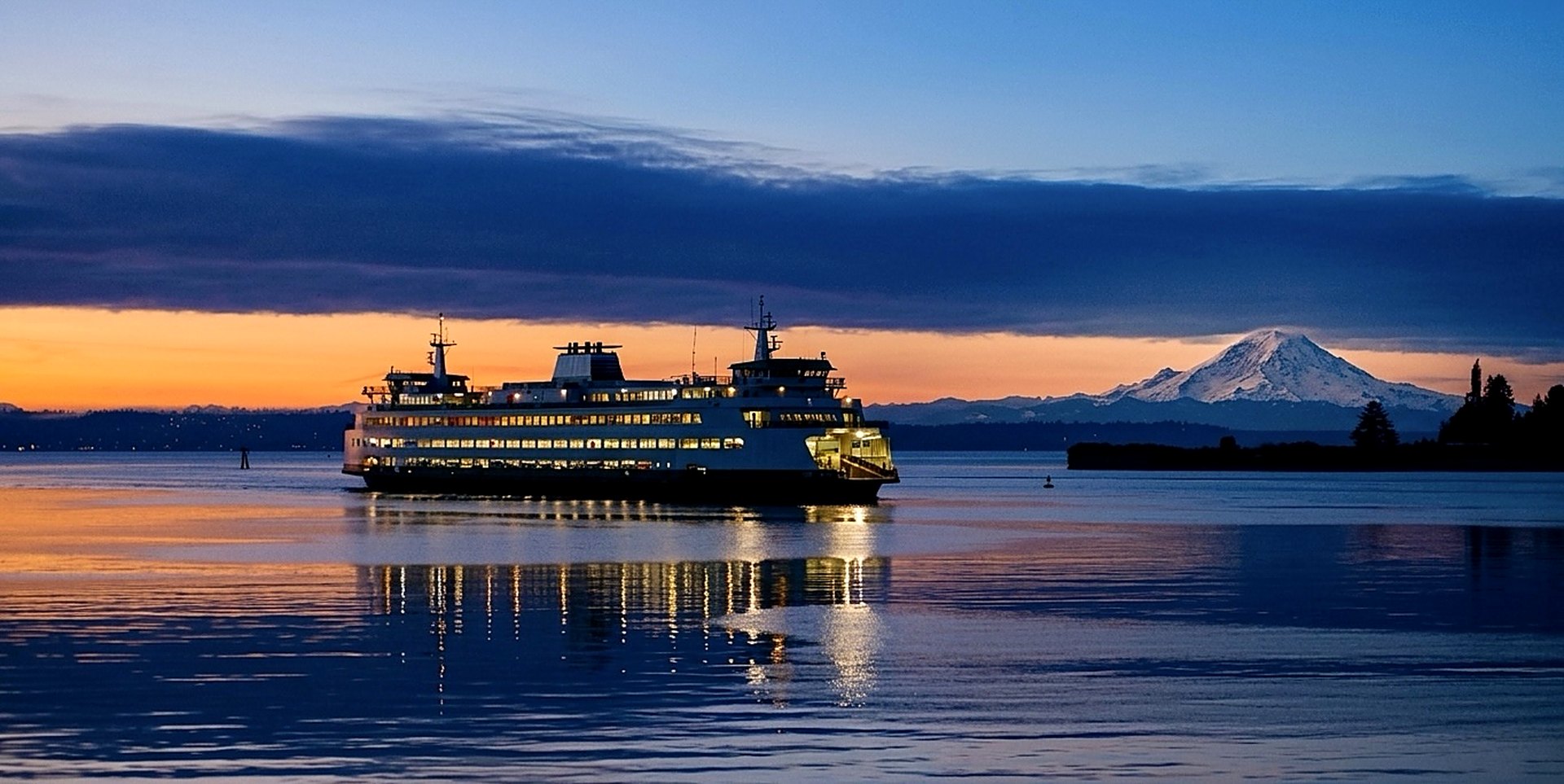 A Washington State Ferry crossing Puget Sound at twilight, its windows glowing warm against the water, with Mount Rainier rising in the distance.